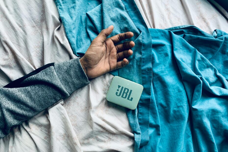 Cozy scene with a hand resting next to a JBL speaker on a blue bedsheet, indoor setting.