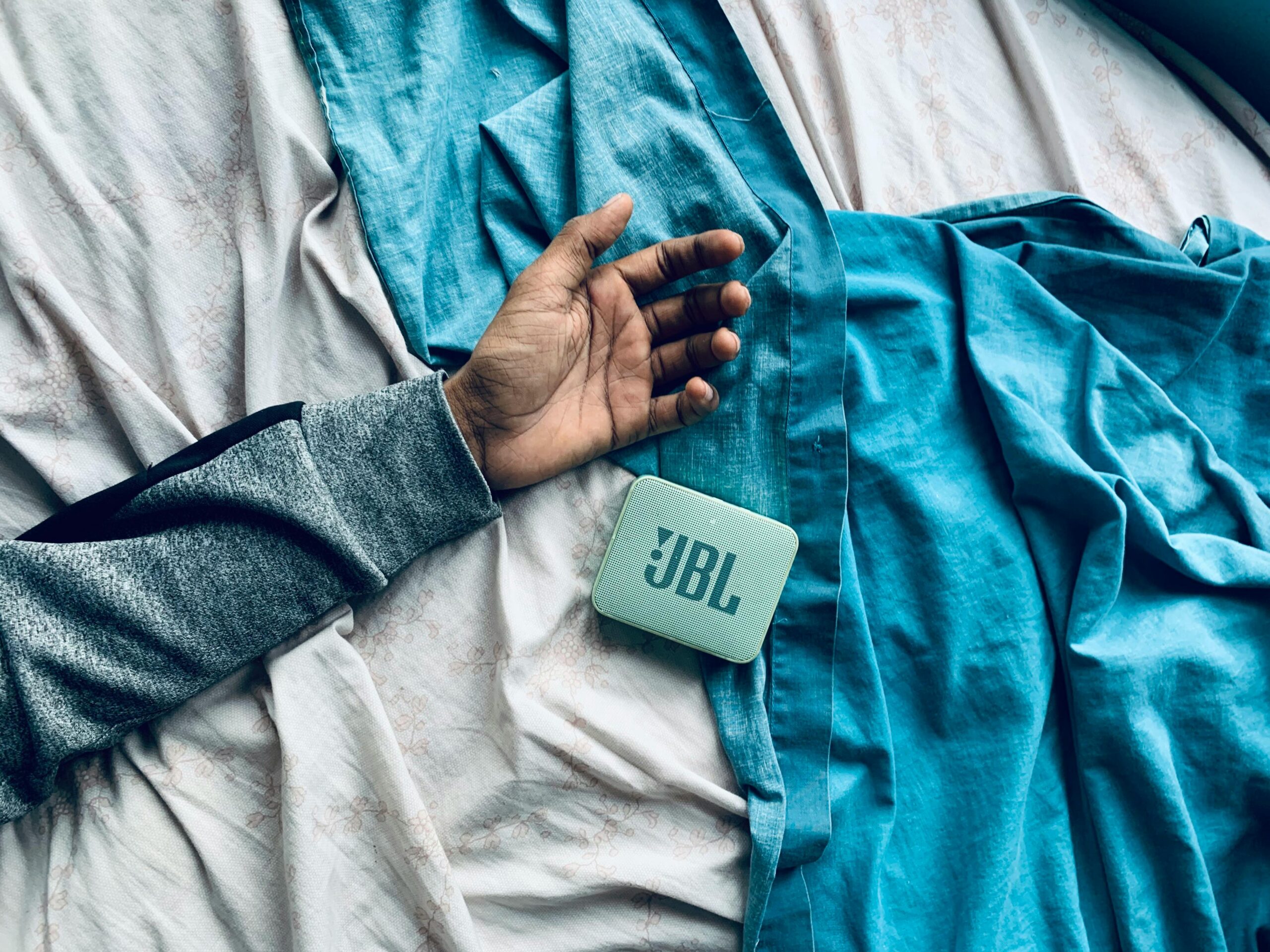 Cozy scene with a hand resting next to a JBL speaker on a blue bedsheet, indoor setting.